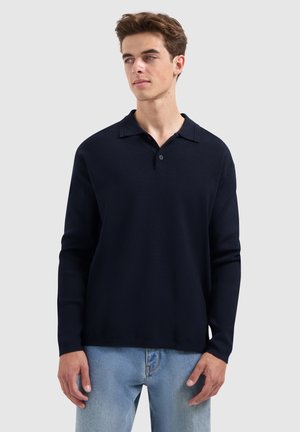 Marineblauwe polo met lange mouwen, vlakke kraag, knoopsluiting met twee knopen, zachte textuur en een comfortabele pasvorm. Gecombineerd met lichtblauwe jeans.