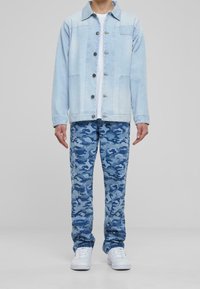 Light blue denim jacket with a button front, paired with blue camouflage pants. White t-shirt underneath, accessorized with white sneakers.