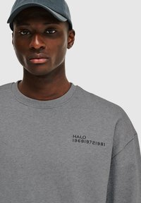 Grijze sweatshirt met een ronde hals en geborduurde tekst "HALO 196819721981" in zwart op de linkerborst. Zachte textuur, casual pasvorm.