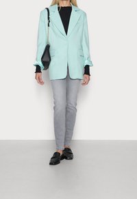 Blazer ajustado verde menta con hombros estructurados, puños de satén negro, combinado con jeans ceñidos gris claro y mocasines negros.
