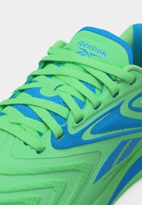 Κοντινό πλάνο ενός φωτεινού πράσινου και μπλε αθλητικού παπουτσιού Reebok, με εστίαση στα κορδόνια, το ύφασμα από δίχτυ και το λογότυπο στη γλώσσα και την πλευρά.