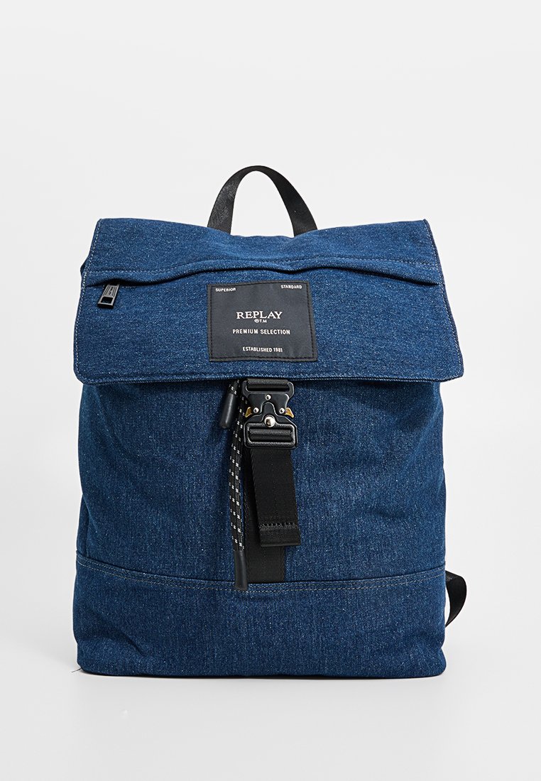 Blauwe denim rugzak met zwarte schouderband en metalen gesp, voorzien van een Replay merkpatch met de tekst "Premium Selection, Established 1981."
