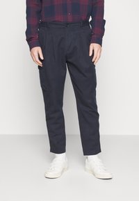 Pantaloni cargo blu navy con pinces, realizzati in tessuto resistente. Presentano tasche laterali, una chiusura con bottone e gambe affusolate. Indossati con sneakers bianche.