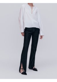 Blouse blanche à manches longues avec col en V et épaules bouffantes, associée à un pantalon en jean foncé avec fentes latérales et des chaussures noires à bouts pointus.