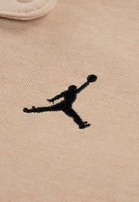 Tecido bege com textura lisa, apresentando um logótipo preto bordado de uma figura a saltar a segurar uma bola de basquetebol.