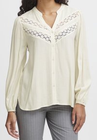 Blouse crème clair avec un col en V, fermeture à boutons, manches longues et dentelle sur le buste avec un motif géométrique.