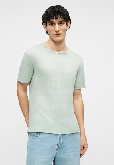 Νέος άνδρας με καστανά σγουρά μαλλιά που φοράει ανοιχτά πράσινο κοντομάνικο πουκάμισο και ανοιχτό μπλε τζιν, στέκεται μπροστά σε απλό φόντο.