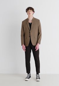Brauner, maßgeschneiderter Blazer über einem schwarzen Top, kombiniert mit schmalen, dunklen Hosen und schwarzen High-Top-Sneakers. Glattes Material mit modernem Schnitt.