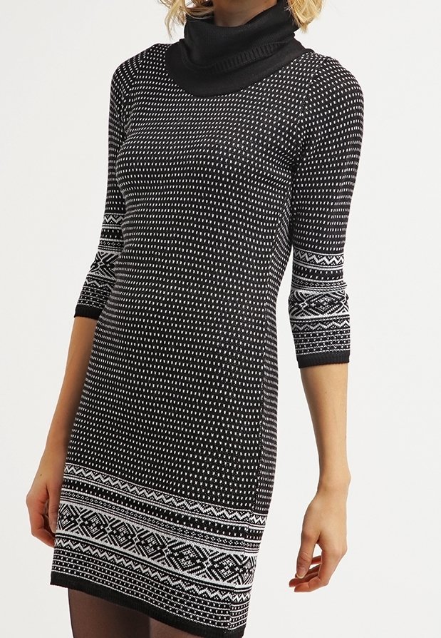 Femme portant une robe tricotée à motif noir et blanc avec des manches trois-quarts et un col roulé noir.
