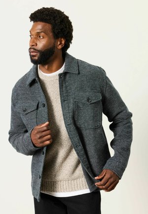 Mann mit grauer Knopfleistenjacke über einem beigefarbenen Strickpullover, richtet die Jacke und blickt nach links vor schlichtem Hintergrund.