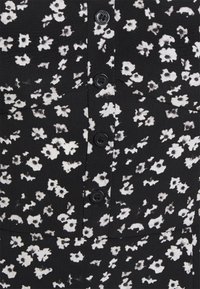 Svart tyg med ett vitt blommönster, med fem svarta knappar fram och två bröstfickor. Mjukt, strukturerat material.