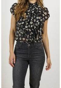 Svart blus med beige blommönster, volangdesign framtill och hög halsringning, kombinerad med mörka, åtsittande jeans. Strukturerad tyg synlig.