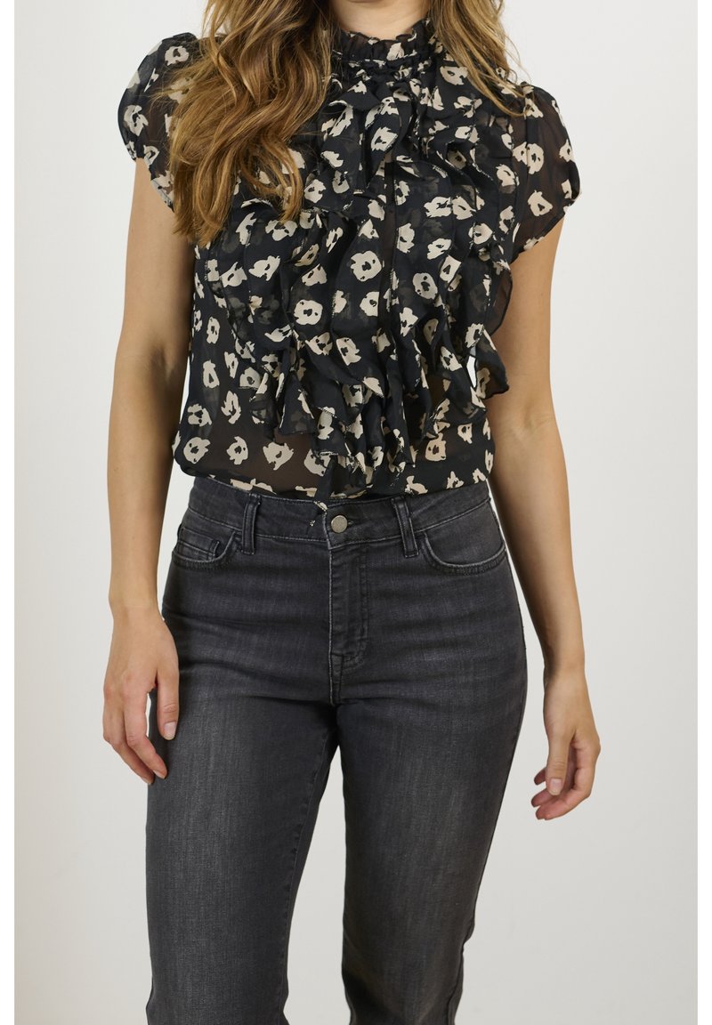 Svart blus med beige blommönster, volangdesign framtill och hög halsringning, kombinerad med mörka, åtsittande jeans. Strukturerad tyg synlig.