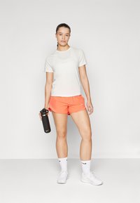 Camisa blanca de manga corta, pantalones cortos naranjas y zapatillas blancas. Sosteniendo una botella de agua negra. Logo de Nike en la camisa, los pantalones cortos y los calcetines.