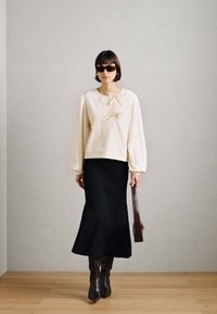 Haut crème à manches longues avec un détail en nœud, associé à une jupe évasée noire. Le mannequin porte des lunettes de soleil noires et des bottes brunes à hauteur de genoux, tout en tenant un sac.