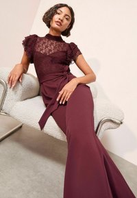 Un vestido burdeos con corpiño de encaje, mangas con volantes y un cinturón atado a la cintura. Tejido suave y fluido, combinado con una silla texturizada de color blanco roto.