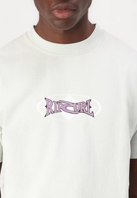 Camiseta de algodón de un suave verde menta con un escote redondo, que presenta un logo "Ripcurl" en color rosa con una fuente estilizada en el centro. Textura suave.
