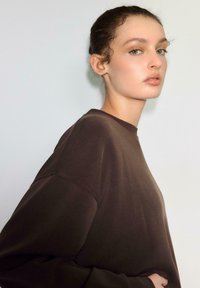 Rjava majica z dolgimi rokavi, sproščenega kroja, z okroglim izrezom in mehkobo teksture; gladek material z minimalnimi šivi.