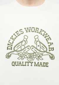 Πράσινο κείμενο "Dickies Workwear Quality Made" με δύο σταυρωμένα αγκίστρια και τρία λουλούδια σε λευκό πουκάμισο.