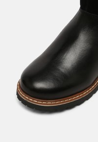Gros plan d'une botte en cuir noire à bout rond, avec une trépointe cousue marron et blanche et une semelle épaisse en caoutchouc noir.