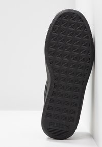Semelle de chaussure Puma noire avec motif étoile géométrique texturé et logo Puma près de la zone du talon, inclinée contre une surface d’angle blanche.