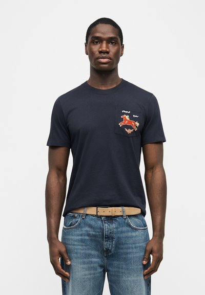 Άνδρας που φοράει ναυτικό μπλε κοντομάνικο με πορτοκαλί κέντημα αλόγου στην τσέπη στο στήθος, ανοιχτό καφέ ζώνη και μπλε τζιν, στέκεται μπροστά σε λευκό φόντο.