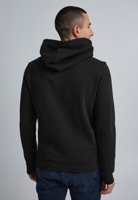 Man in een zwarte hoodie en blauwe jeans, met zijn rug naar de kijker, tegen een effen grijze achtergrond.