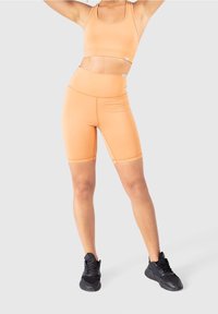 Oranžová sportovní podprsenka a odpovídající vysoké šortky vyrobené z elastické látky. Černé sportovní boty. Hladká textura, přiléhavý design.