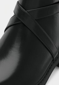 Čierne členkové topánky vyrobené z lesklej kože, s krížovými popruhmi a zaoblenou špičkou. Hladký povrch s šitými akcentmi pozdĺž podrážky.