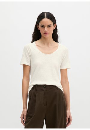 Frau mit langen dunklen Haaren, die ein weißes kurzärmeliges, rundhalsausschnitt Shirt und eine hoch taillierte dunkelbraune Hose trägt, stehend vor einem einfarbigen Hintergrund.