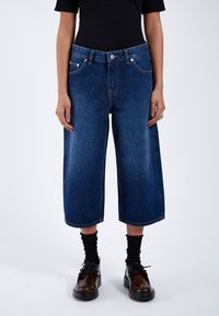 Tummansiniset denim-culottes leveällä lahkeella, näkyvillä etutaskuilla ja nappikiinnityksellä. Yhdistetty mustiin ribbikankaisiin sukkiin ja kenkiin.