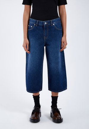 Pantalones culottes de mezclilla azul oscuro con un diseño de pierna ancha, bolsillos frontales visibles y cierre de botón. Combinados con calcetines negros de canalé y zapatos.