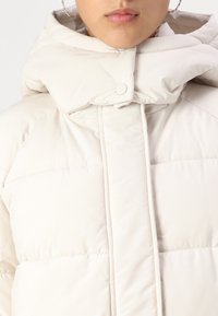 Veste blanche rembourrée avec un col montant, fermeture à boutons-pression et texture matelassée. Tissu doux avec une finition lisse.