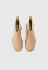 Botine din suede bej cu o textură netedă, panouri elastice laterale și o buclă de prindere. Detalii cusute pe talpa exterioară.