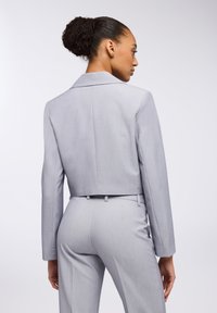 Giacca da abito grigia chiara cropped con colletto a punta e maniche lunghe. Vestibilità sartoriale con superficie liscia, abbinata a pantaloni slim coordinati.