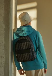 Rucsac negru cu model geometric, cu un fund neted negru, purtat pe un hanorac teal, asortat cu o pălărie bej de tip bucket.