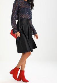 Blouse marine à motif rouge, jupe plissée en cuir noir et bottines en daim rouge. Un petit sac bicolore complète le look.