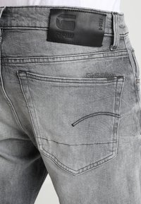Jeans de mezclilla gris con un parche de cuero etiquetado como "RAW" en la cintura. Presentan bolsillos traseros estándar y un detalle de costura curva.