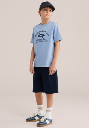 Băiat purtând un tricou cu imprimeu albastru deschis, pantaloni scurți bleumarin, șosete în dungi, adidași albaștri și o șapcă închisă la culoare, stând într-un fundal simplu.