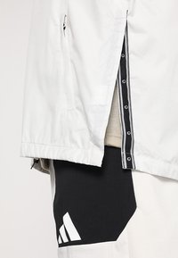 Veste gris clair avec une poche latérale zippée, un accent noir et blanc à l'intérieur, et un tissu noir contrastant en dessous avec un détail logo.
