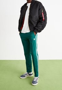 Svart bomberjacka med en dragkedjeficka, vit t-shirt, gröna träningsoverallbyxor med vita ränder och grå sneakers. Släthet i texturen, avslappnad design.