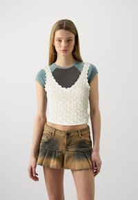 Fehér horgolt crop top, fodros nyakkivágással, szürke pólóra rétegezve. Barna farmer szoknyával párosítva, amelynek bővülő alja van.