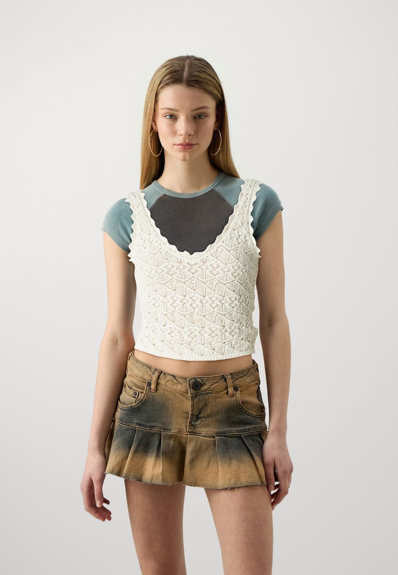 Fehér horgolt crop top, fodros nyakkivágással, szürke pólóra rétegezve. Barna farmer szoknyával párosítva, amelynek bővülő alja van.