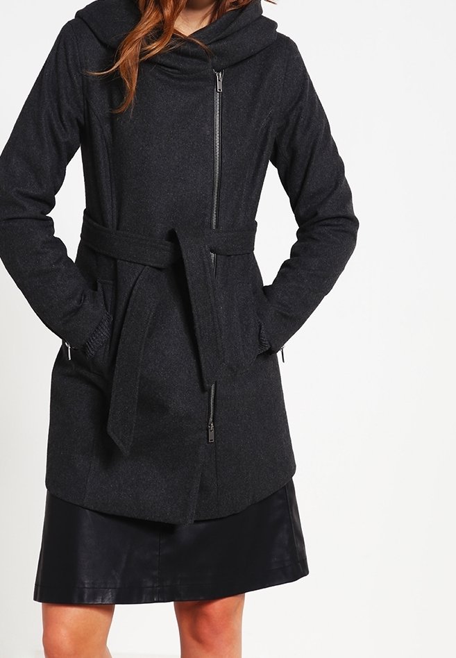 Abrigo negro hecho de tela texturizada, con cuello tipo embudo, cinturón en la cintura, mangas largas y un bolsillo delantero con cremallera. Llevado parcialmente sobre una falda de cuero.