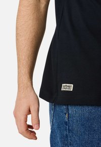 Black cotton t-shirt with a straight cut, short sleeves, and a visible logo tag at the hem, paired with blue denim jeans.