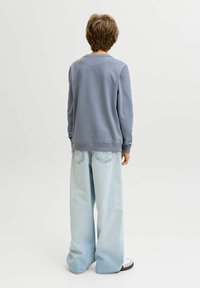 Παιδί με ανοιχτό καστανά μαλλιά στέκεται γυρισμένο με την πλάτη, φοράει φαρδιά μπλε φούτερ, φαρδιά ανοιχτόχρωμα τζιν και άσπρα αθλητικά παπούτσια σε ένα απλό φόντο.