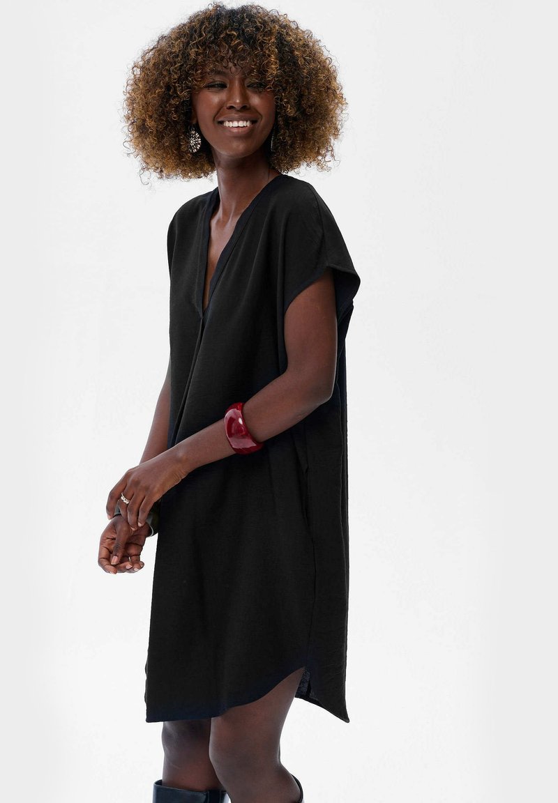 Czarna sukienka krótki rękaw z dekoltem w kształcie V, luźny krój i boczne rozporki. Na nadgarstku noszony czerwony bransoletka. Teksturowany materiał i swobodny kształt.