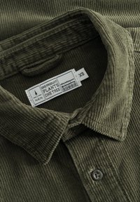 Chemise en velours côtelé vert olive avec un col rabattu, deux boutons gris et une étiquette indiquant "NIKIN" et "XS" à l'intérieur du col.