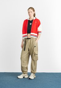 Cardigã curto em vermelho e branco com acabamento riscado, top cropped em malha preta, calças cargo bege com bolsos, e tênis claros. O fundo é branco.