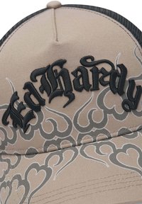 Casquette beige avec dos en mesh noir, présentant un texte brodé noir "Bad Hardy" et des motifs de cœur gris en forme de flammes sur le devant.
